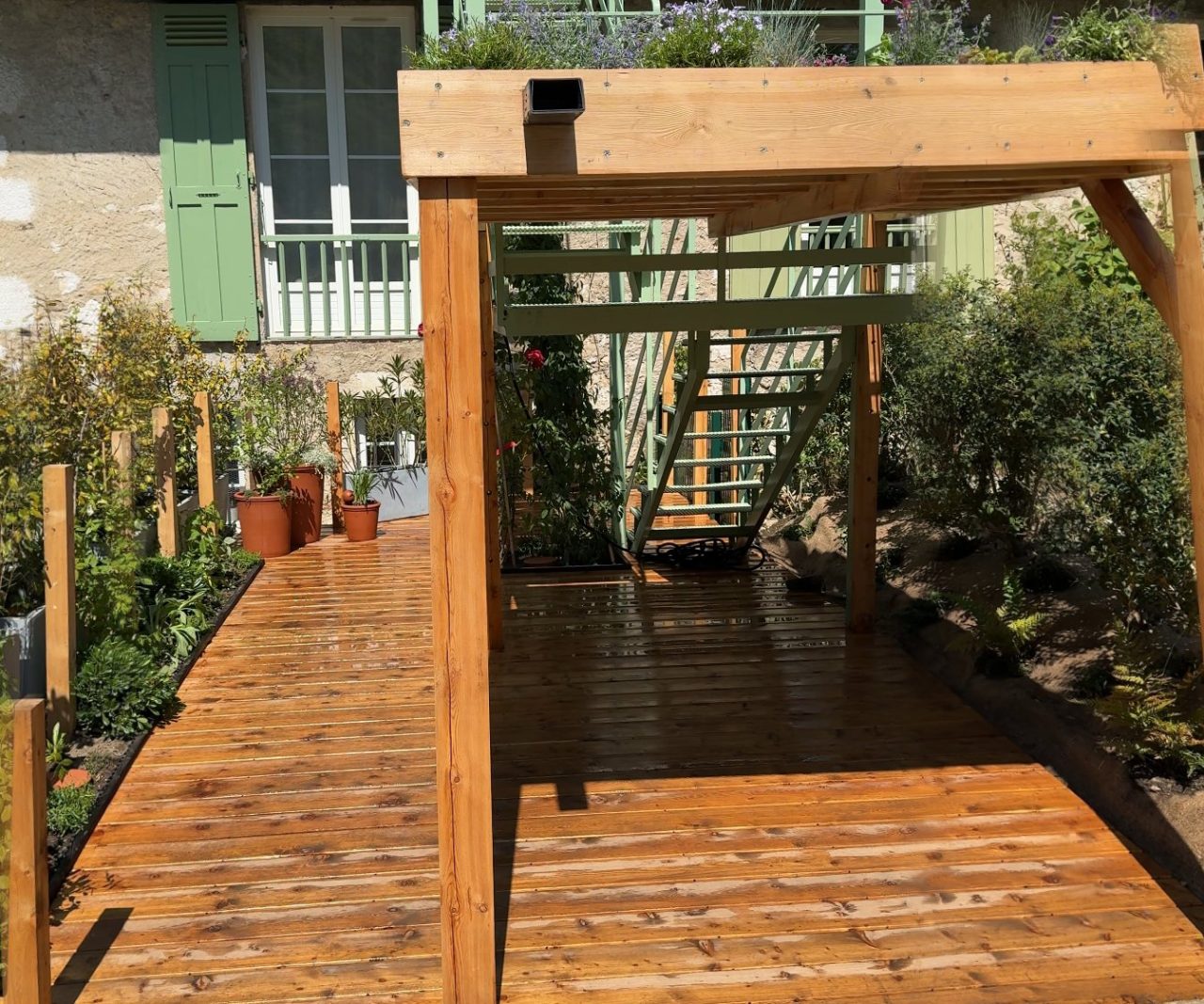 La même cour après les travaux : terrasse en mélèze, pergola à toiture végétale, plantations et portillon